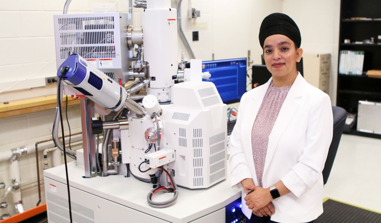 Jasneet Kaur, wearing a white lab coat, stands next to a large electron microscope in a science lab at Brock University.