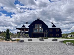 River House Winery, a large blue barn-like building, sits atop a small hill and against a blue sky with clouds.