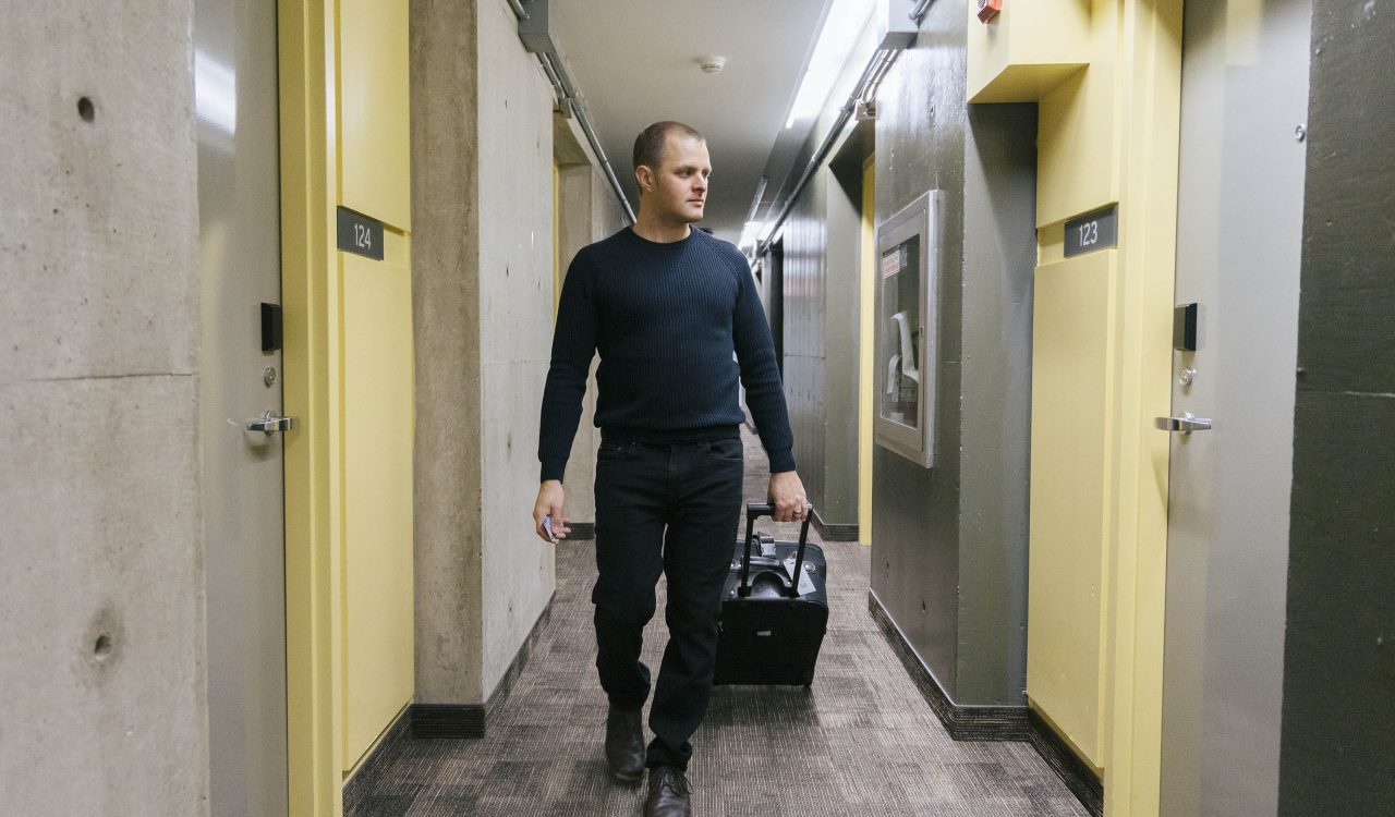 A man walks down a hallway pulling a suitcase on wheels.