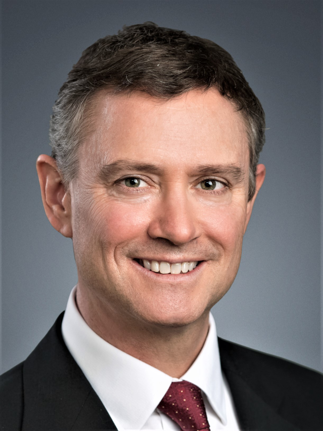 Simon Haynes poses wearing a dark suit, white dress shirt and red tie on a solid grey background.