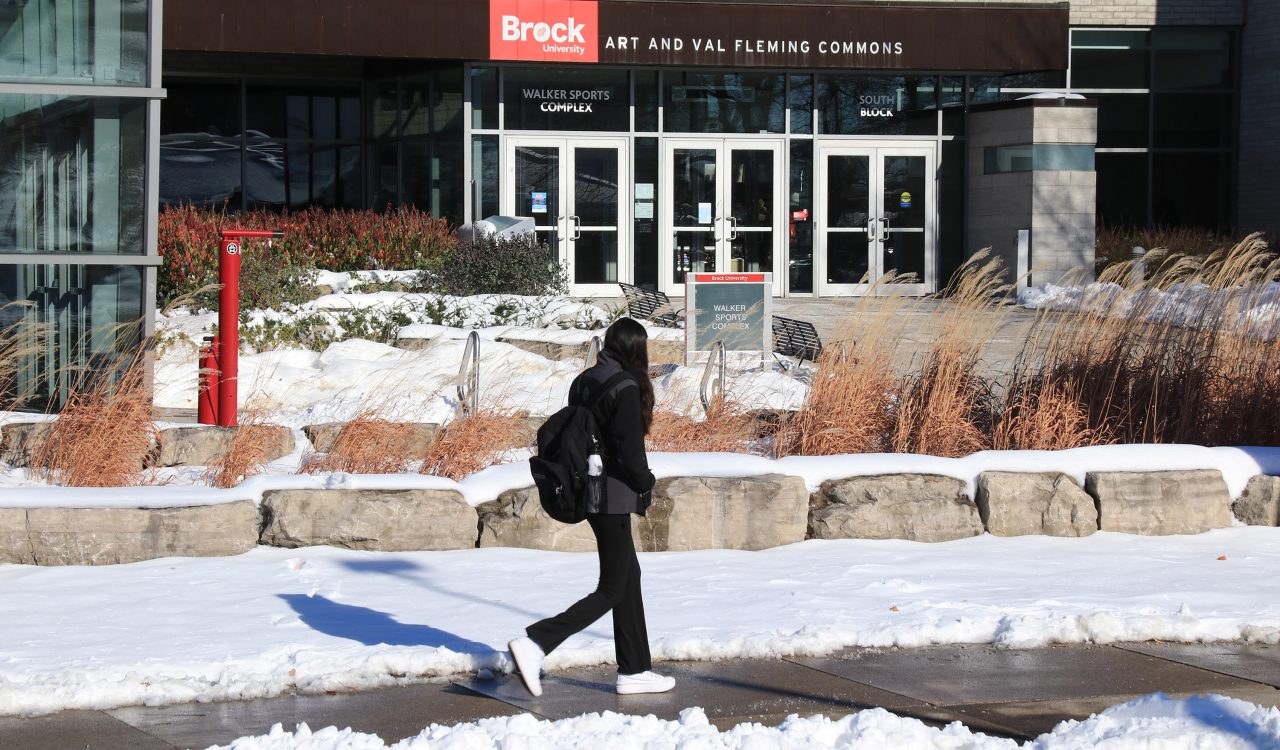 A Brock University student walks on the sidewalk in front of the Art and Val Fleming Commons. Snow covers the lawn and bushes around her.