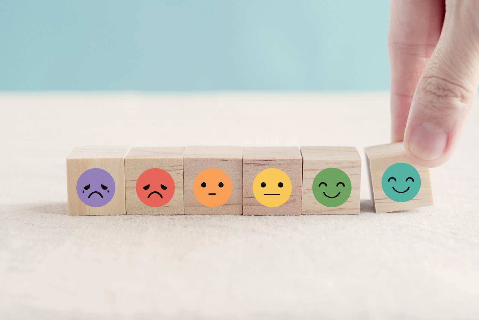Closeup of small wooden blocks, each with a cartoon face expressing a different emotion, from very sad to neutral to very happy. A hand is reaching down, placing the very happy block on the table.