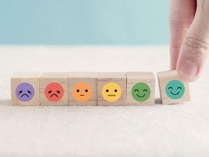 Closeup of small wooden blocks, each with a cartoon face expressing a different emotion, from very sad to neutral to very happy. A hand is reaching down, placing the very happy block on the table.