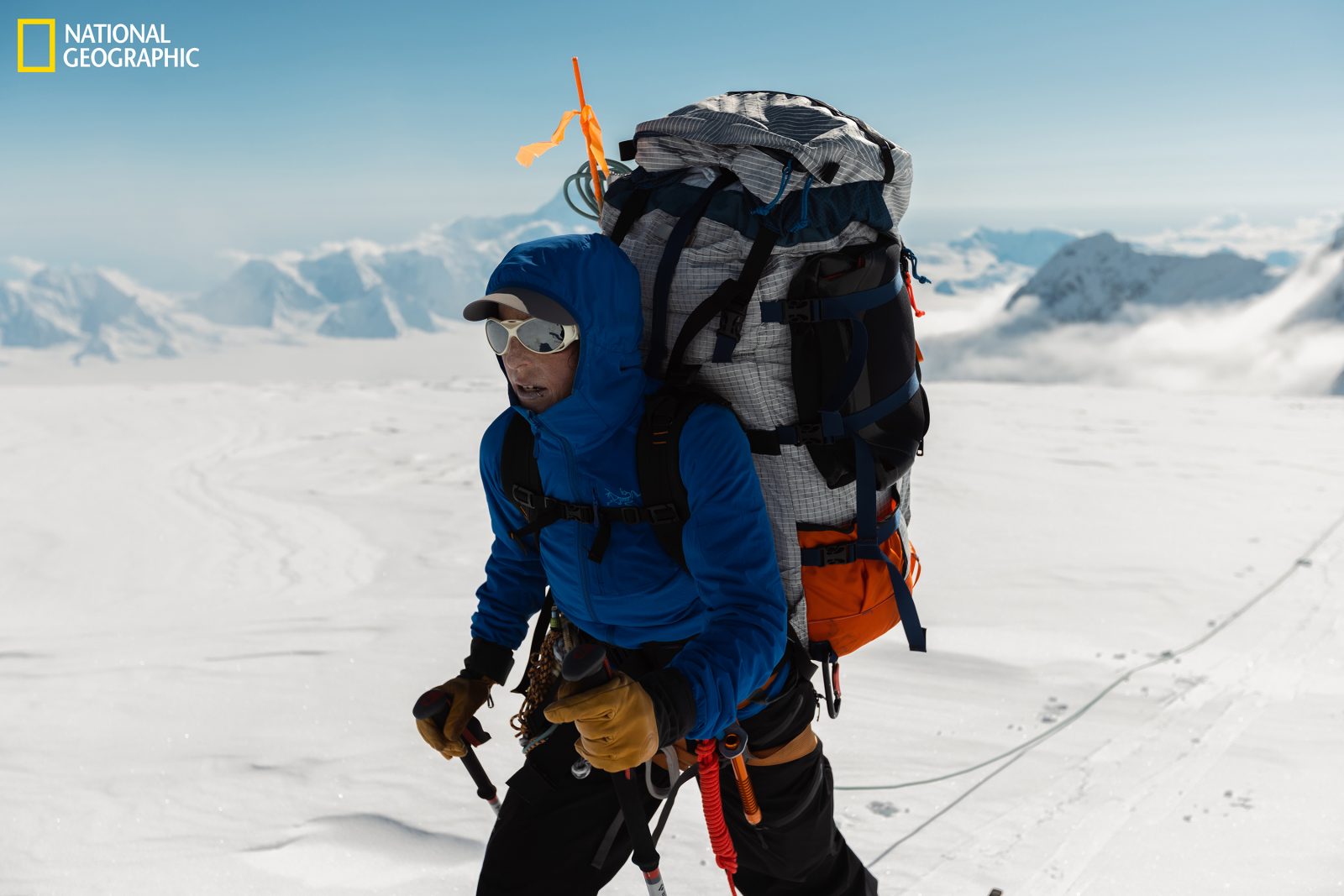 Alison Criscitiello wears a blue mountaineering jacket and gear-filled hiking backpack while traversing snow-covered flatland at high altitudes on Mount Logan in the Canadian Yukon. She is wearing large sunglasses and using hiking poles while attached to a guide wire. The backdrop is a clear blue sky and massive mountain peaks.