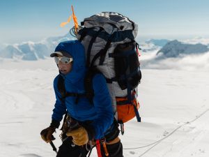 Alison Criscitiello wears a blue mountaineering jacket and gear-filled hiking backpack while traversing snow-covered flatland at high altitudes on Mount Logan in the Canadian Yukon. She is wearing large sunglasses and using hiking poles while attached to a guide wire. The backdrop is a clear blue sky and massive mountain peaks.