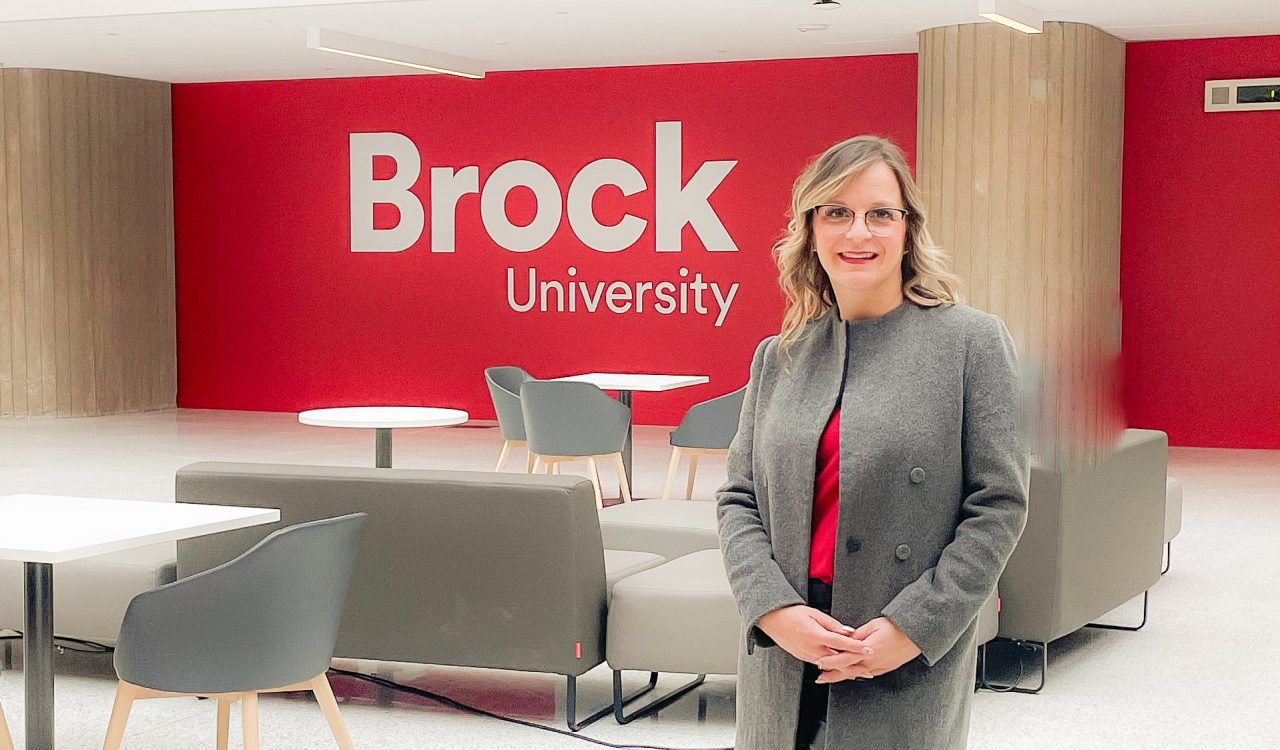 A woman stands in an indoor atrium in front of a red well with a Brock University logo.