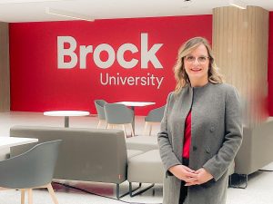A woman stands in an indoor atrium in front of a red well with a Brock University logo.