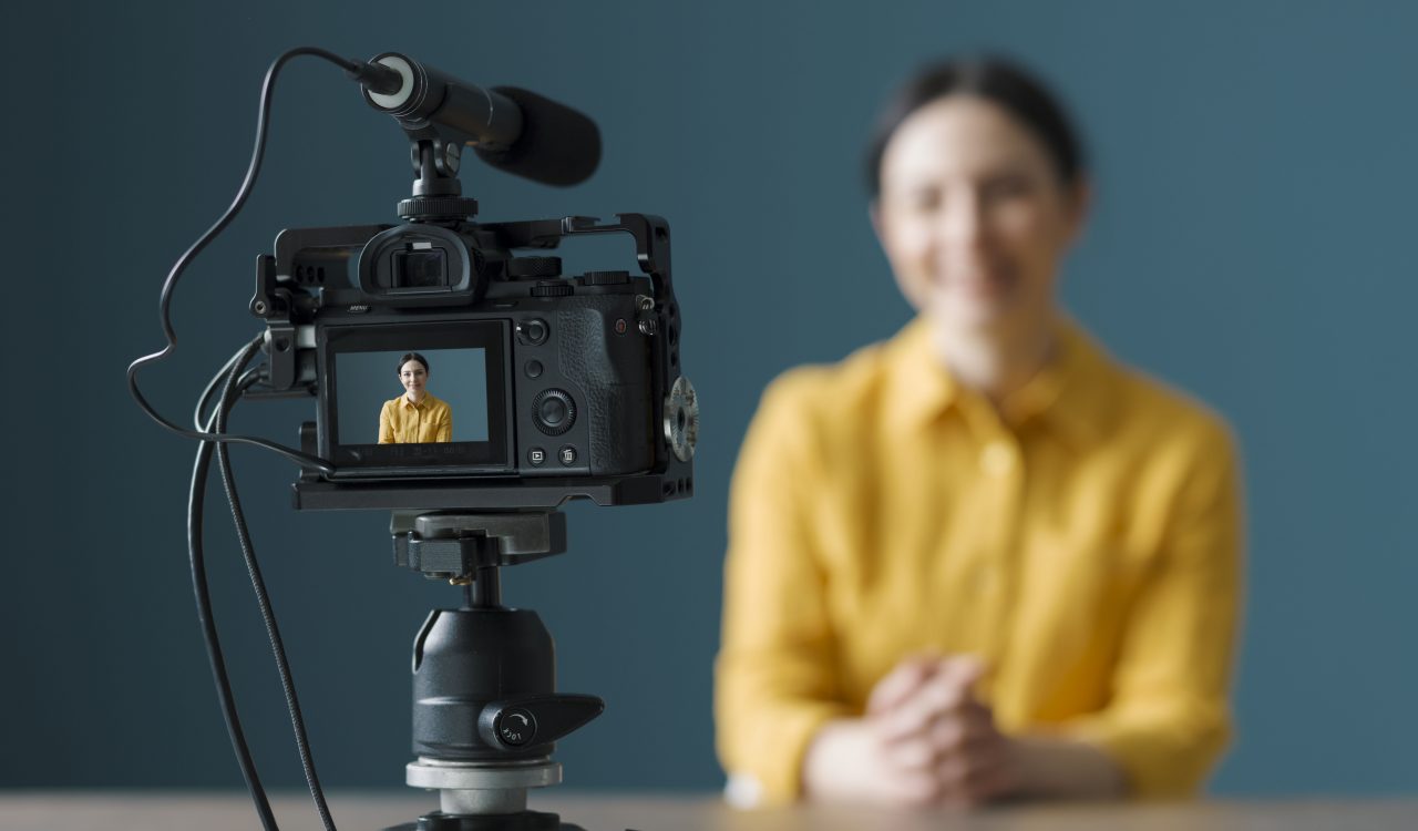 A woman sits in front a camera with a green wall behind her.