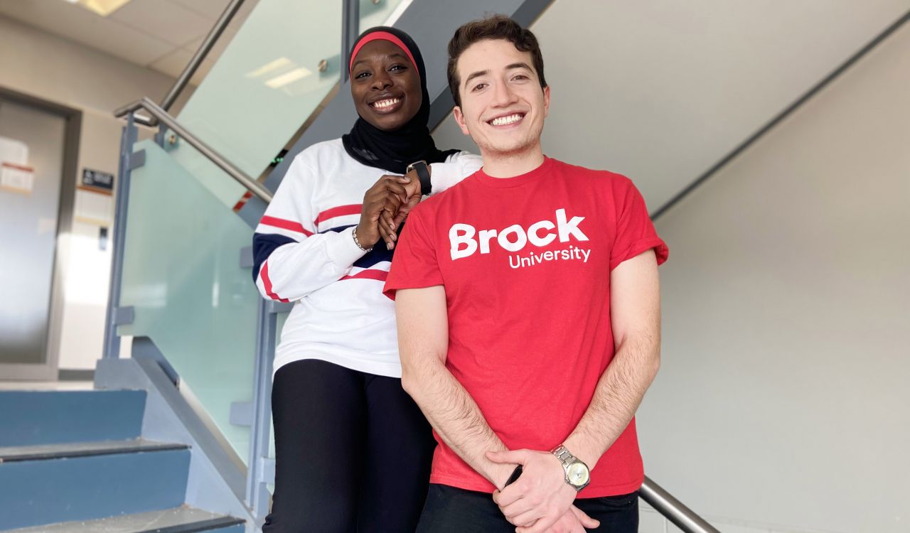 Aishah Sonekan stands behind Sebastian Roa Goyes, resting an arm on his shoulder, both looking towards the camera and smiling in a stairwell in the Brock International office building.