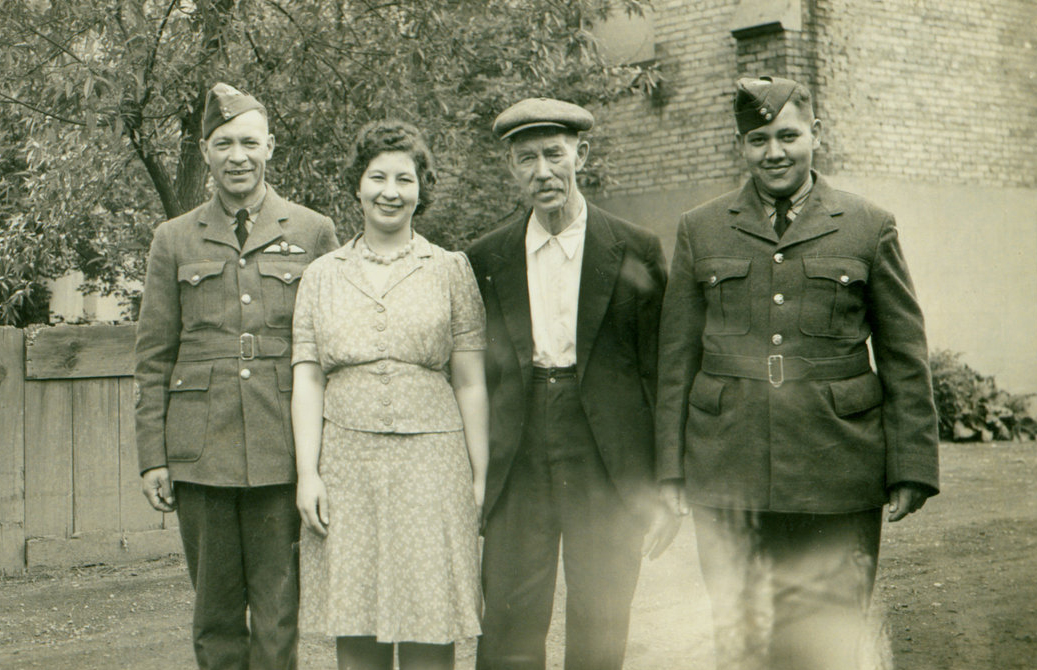 An old sepia coloured photo of four people standing in a row. Two men are wearing military uniforms.