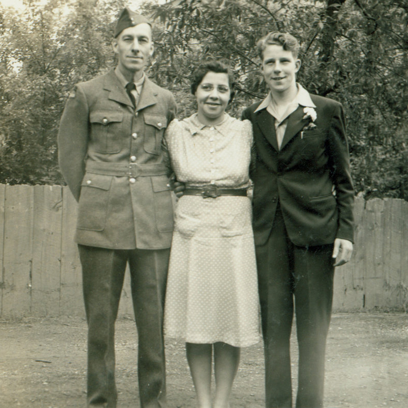 An old sepia coloured photo of three people standing next to each other. A man in a military uniform, a woman in a light-coloured dress and a man in a suit and dress shirt.