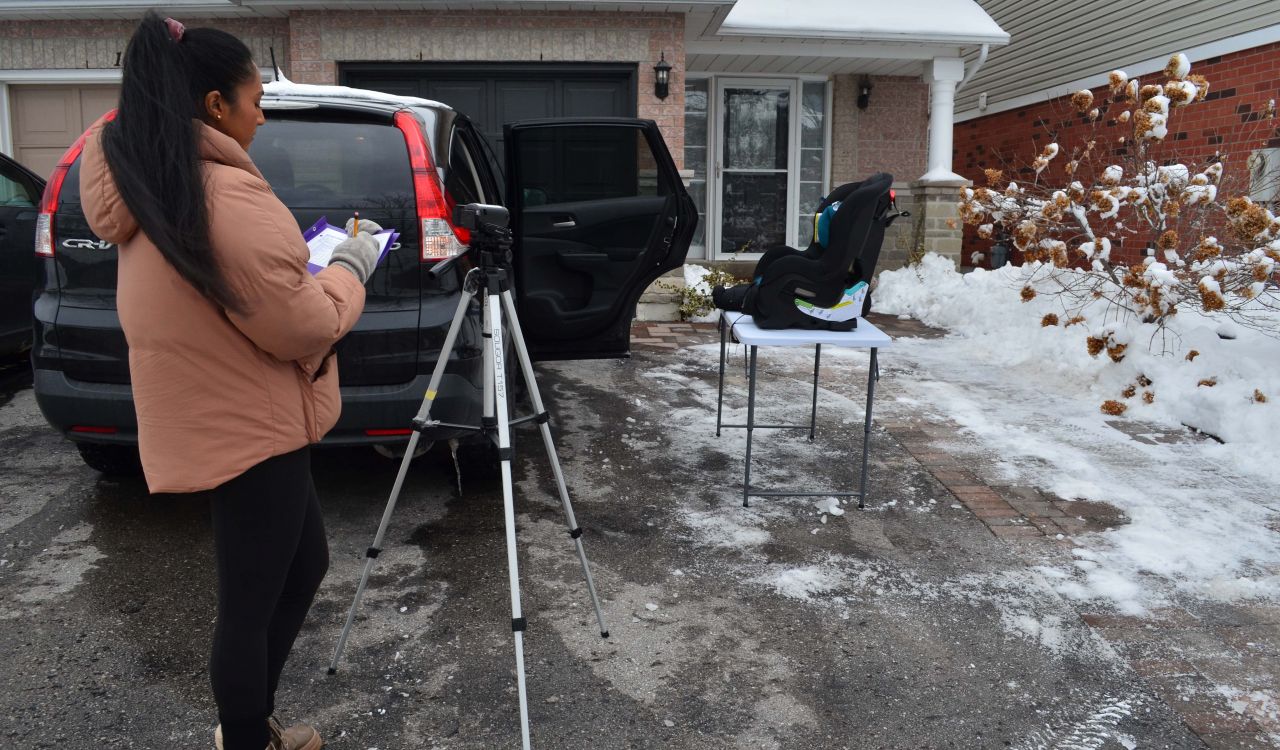 A woman stands outside with a clipboard in hand and a video camera on a tripod in front of her. She looks over towards an SUV and a table with a car seat on it.