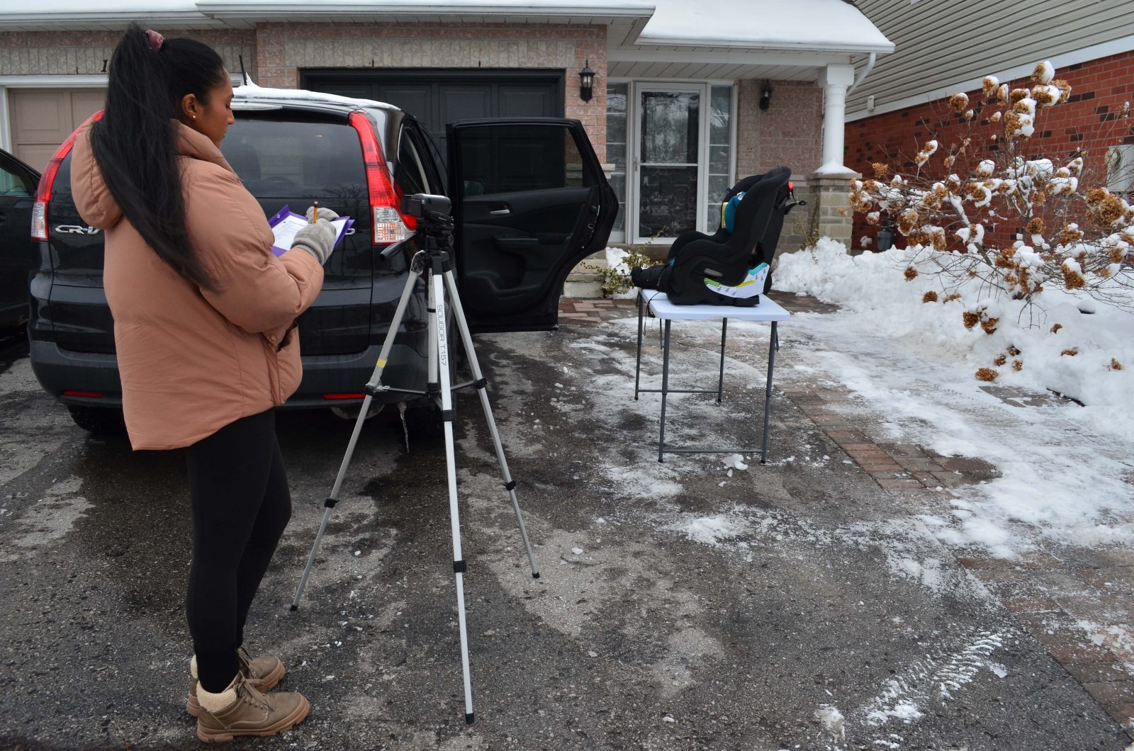 A woman stands outside with a clipboard in hand and a video camera on a tripod in front of her. She looks over towards an SUV and a table with a car seat on it.