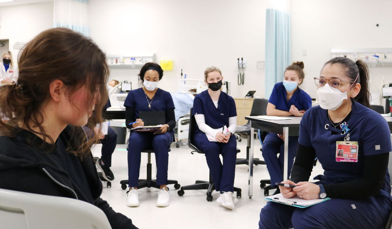 Nursing student Vinita Hatanaka Chotai sits wearing scrubs facing another woman in plain clothing in a clinical setting as several seated Nursing students look on.