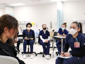 Nursing student Vinita Hatanaka Chotai sits wearing scrubs facing another woman in plain clothing in a clinical setting as several seated Nursing students look on.