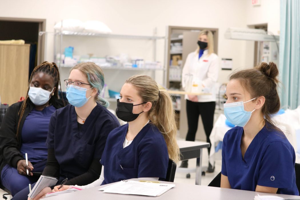 Bachelor of Nursing/Master of Nursing students sit at a table looking in the same direction while a professor stands looking on in the background.