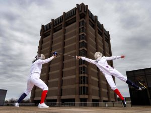 Two women in fencing uniforms battle each other at the base of a university tower.