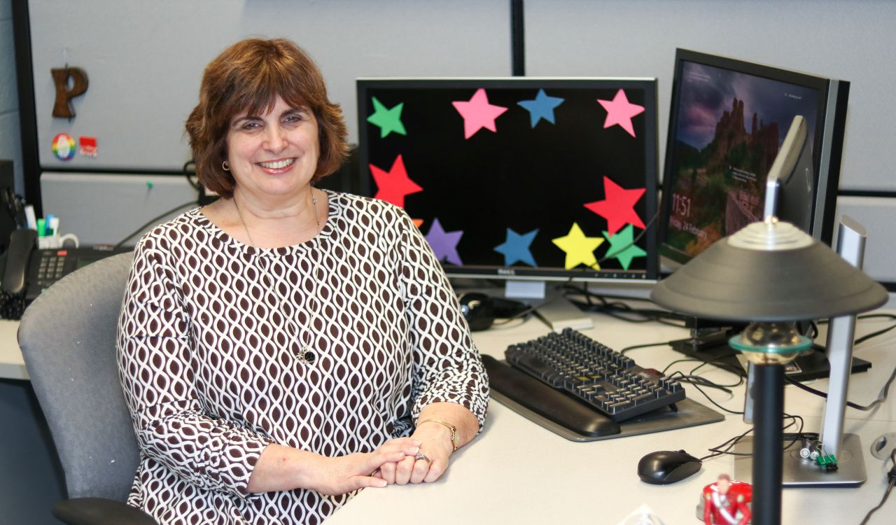 Patricia McDonnell sits at her desk, which has a desktop computer with two screens. Colourful paper cutout stars adorn the perimeter of one of the screens.