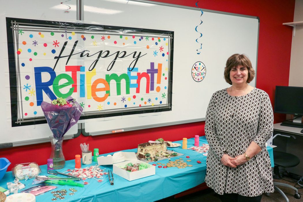 Patricia McDonnell stands in front of a long rectangular table filled with food. A sign on the wall behind her reads “Happy Retirement!”