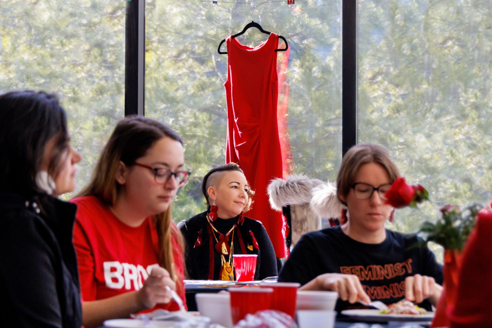 Women sit at a series of tables while a red dress hangs in a window in the background.