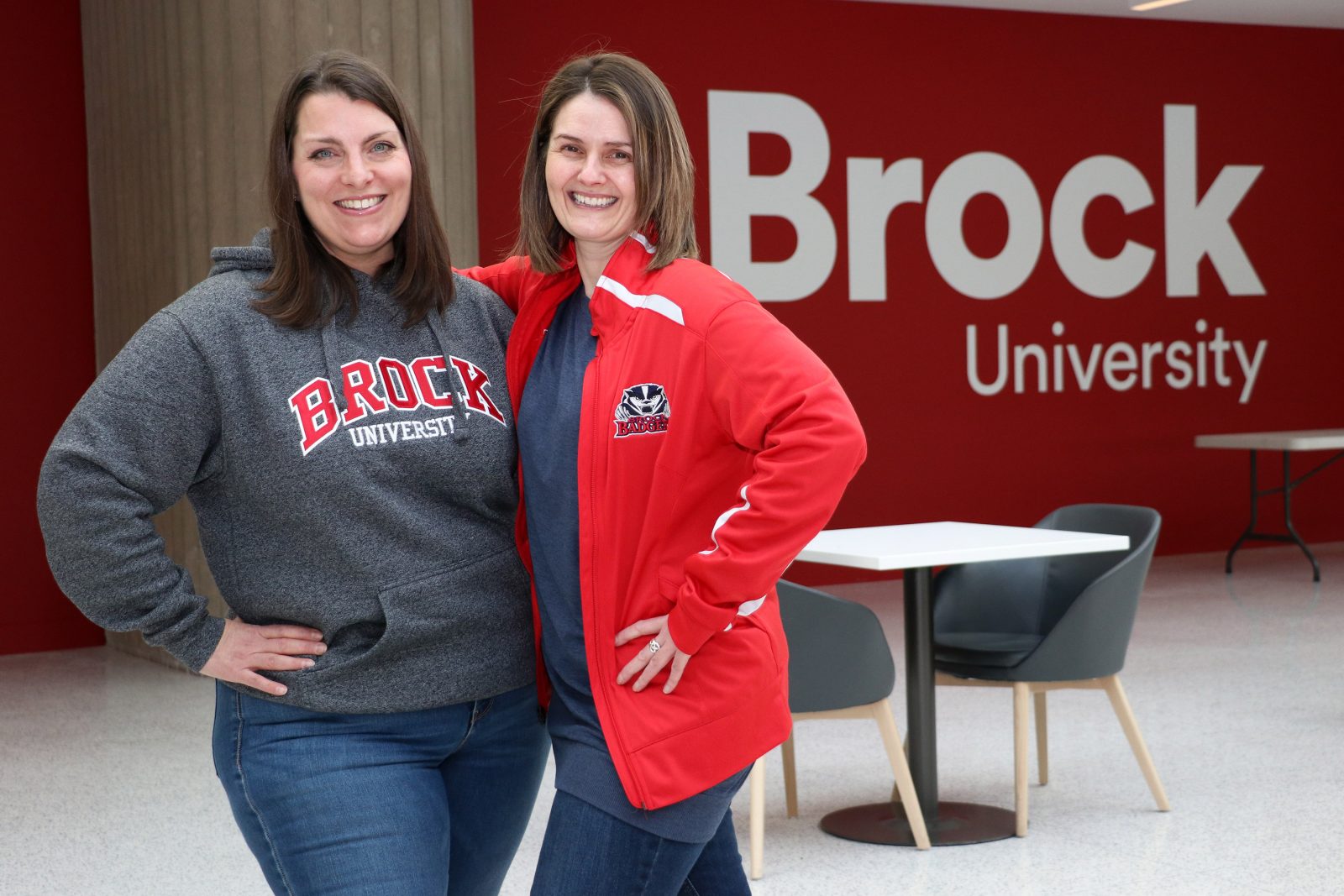Two women pose for a photo in front of a red wall that reads Brock University. Both women are wearing Brock University sweaters.