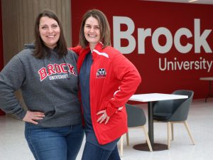 Two women pose for a photo in front of a red wall that reads Brock University. Both women are wearing Brock University sweaters.