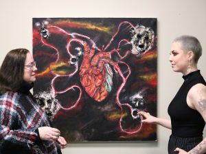 Brock researcher Sheila O’Keefe-McCarthy (left) discusses a mixed-media oil painting of an anatomical heart with 3D faces at the end of four different pictorial representations of veins and arteries extending from the heart with the artist and second-year Brock Nursing student Hannah Michaelson.