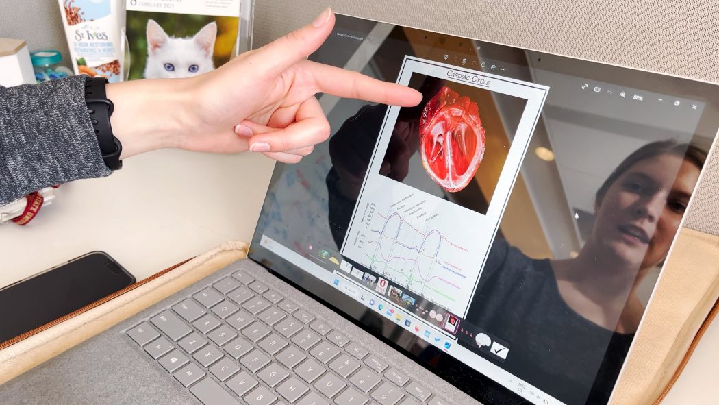 A woman points to an image of an anatomical heart on a computer screen, her reflection visible on the screen.