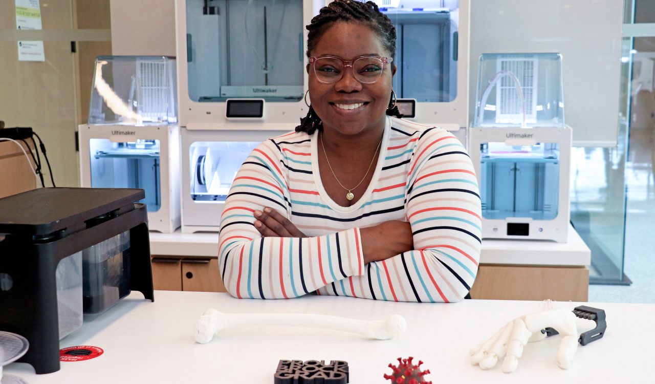 Tabitha Lewis leans on a table in the Brock University Makerspace that displays 3D-printed items. A 3D printer is behind her.