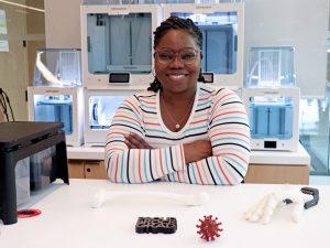 Tabitha Lewis leans on a table in the Brock University Makerspace that displays 3D-printed items. A 3D printer is behind her.