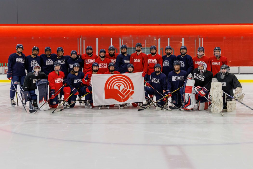 The Brock University Badgers women’s hockey team poses on a sheet of ice holding the United Way flag.
