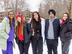 A group of five people stand together outside with snow on the ground and trees in the background.