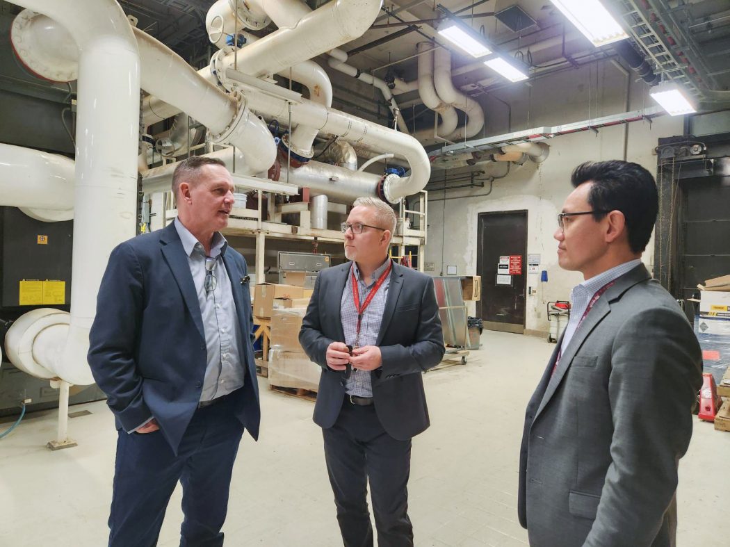 Three men stand speaking to one another in an industrial basement-like setting with large pipes in the background.