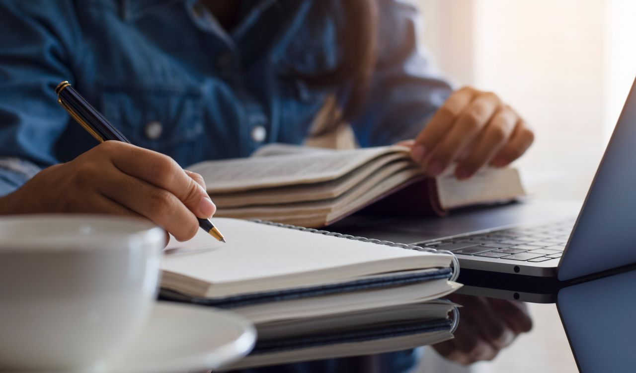 Close up image of a student reading a textbook while also making notes in a notebook. A laptop sits on a table in front of her and a cup of coffee is in the foreground.