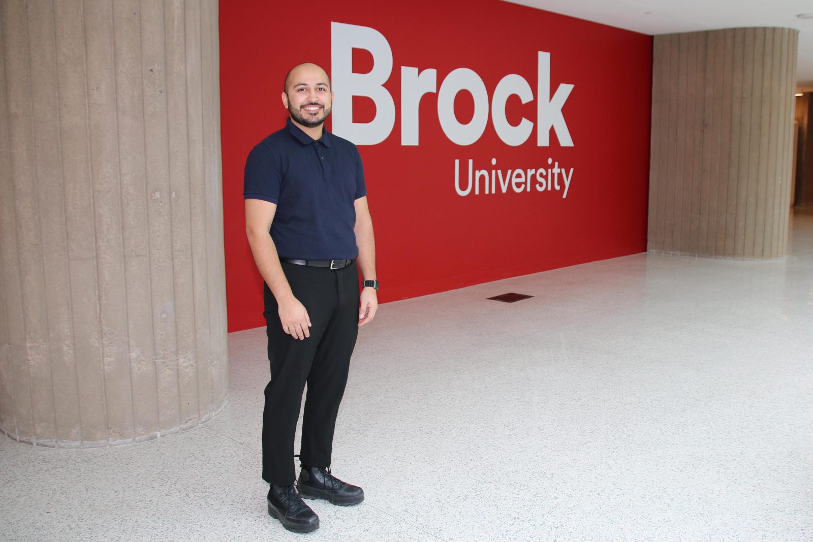 A man stands in front of a large red wall in an indoor atrium that says ‘Brock University.’