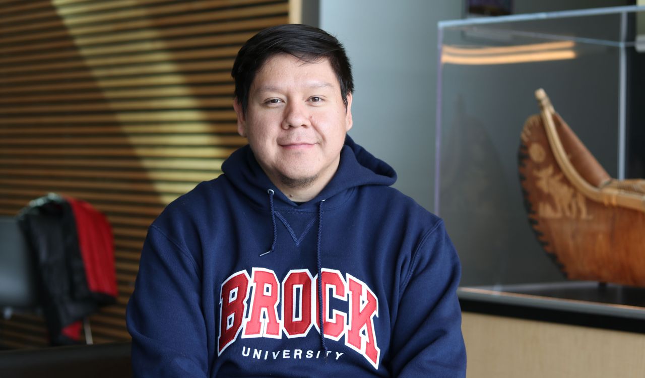 A man in a Brock University shirts sits in an indoor atrium.