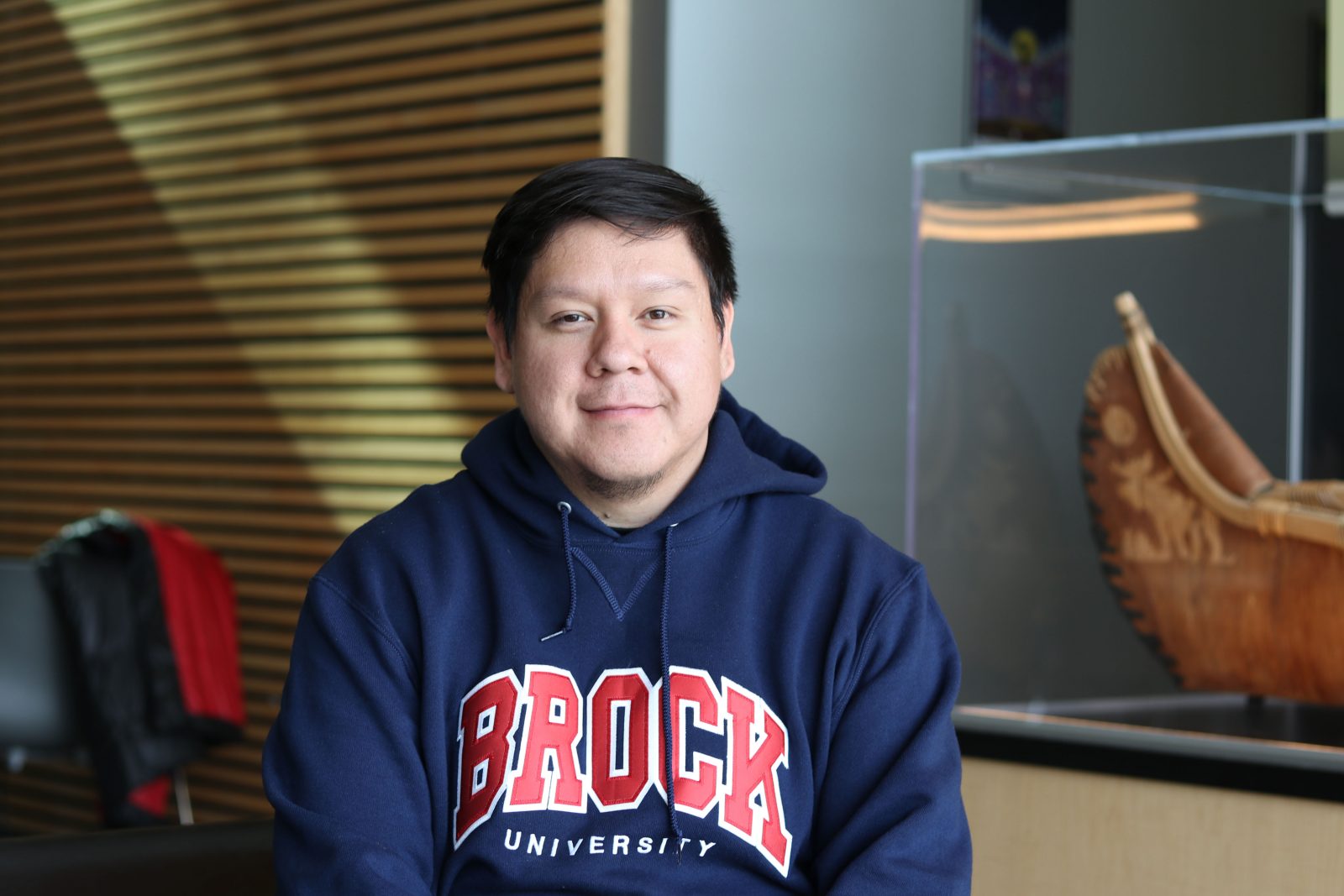 A man in a Brock University shirts sits in an indoor atrium.