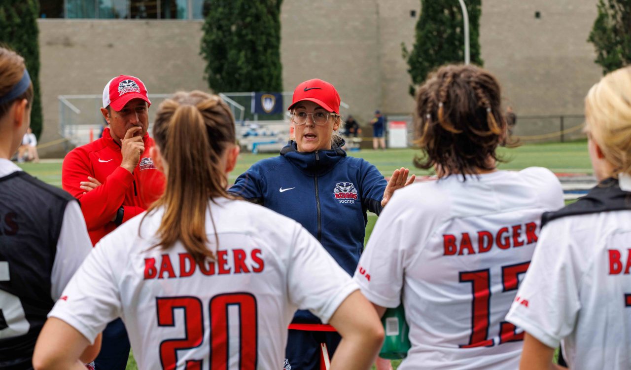 A coach stands speaking to a team of student-athletes on an outdoor field.