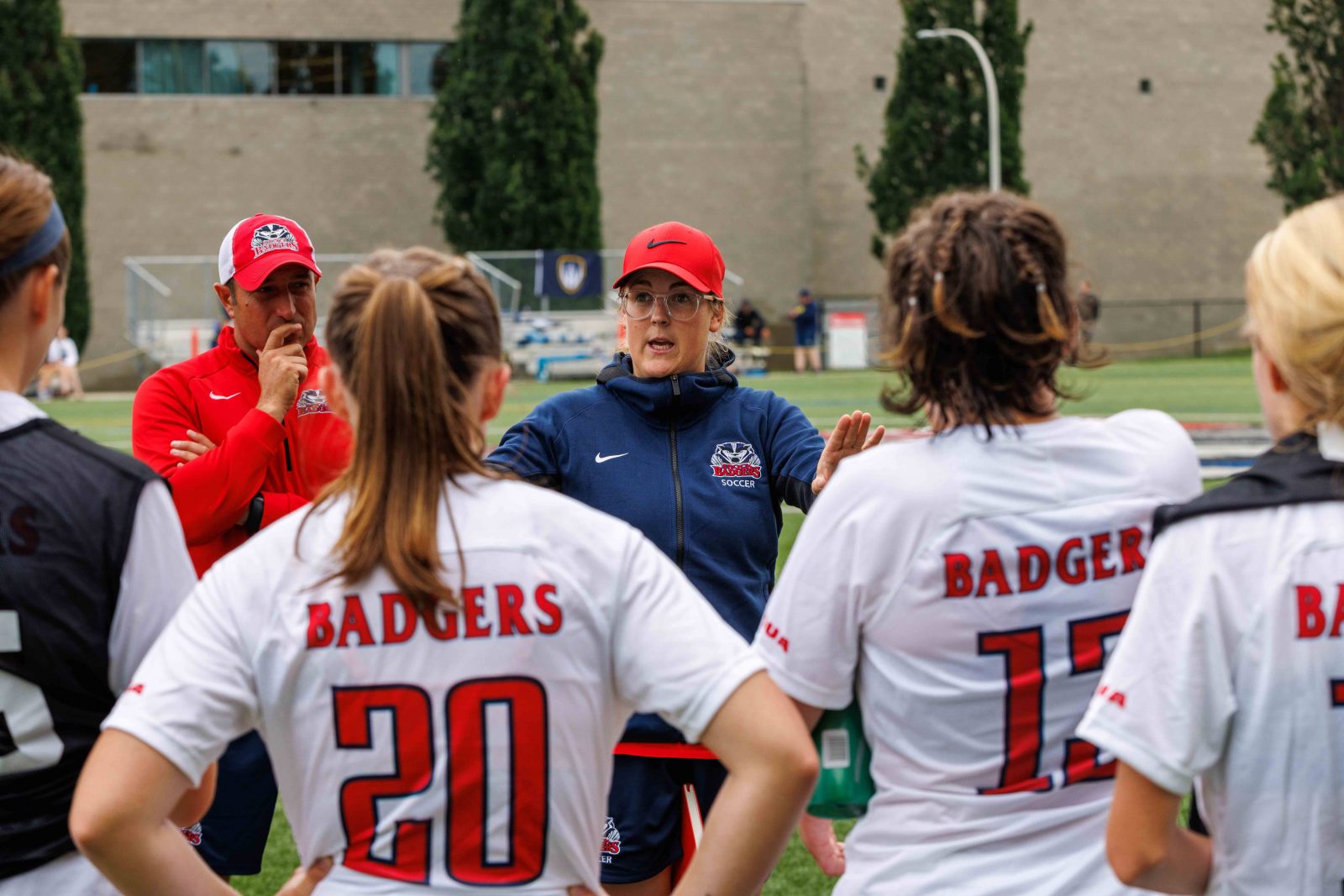 A coach stands speaking to a team of student-athletes on an outdoor field.