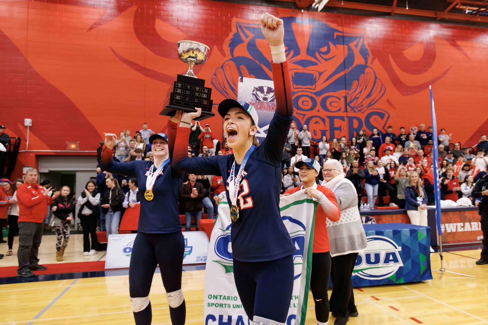 Two women’s volleyball athletes cheer as they hoist a sports trophy surrounded by fans inside a gymnasium.