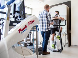 A man collects research data from another man sitting on a stationary bike.
