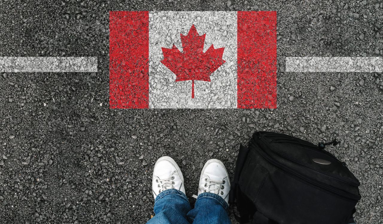 a man with a shoes and backpack is standing on asphalt next to flag of Canada and border