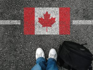 a man with a shoes and backpack is standing on asphalt next to flag of Canada and border