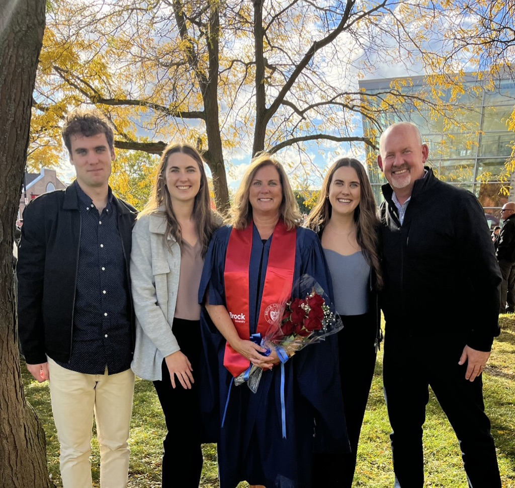 Brock University graduate Kerry Shoalts wears a Convocation robe and stole. She is holding a bouquet of flowers and is flanked by two family members on each side of her. 