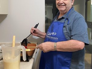 A older man wearing an apron holds a spatula over a grill as he makes pancakes.