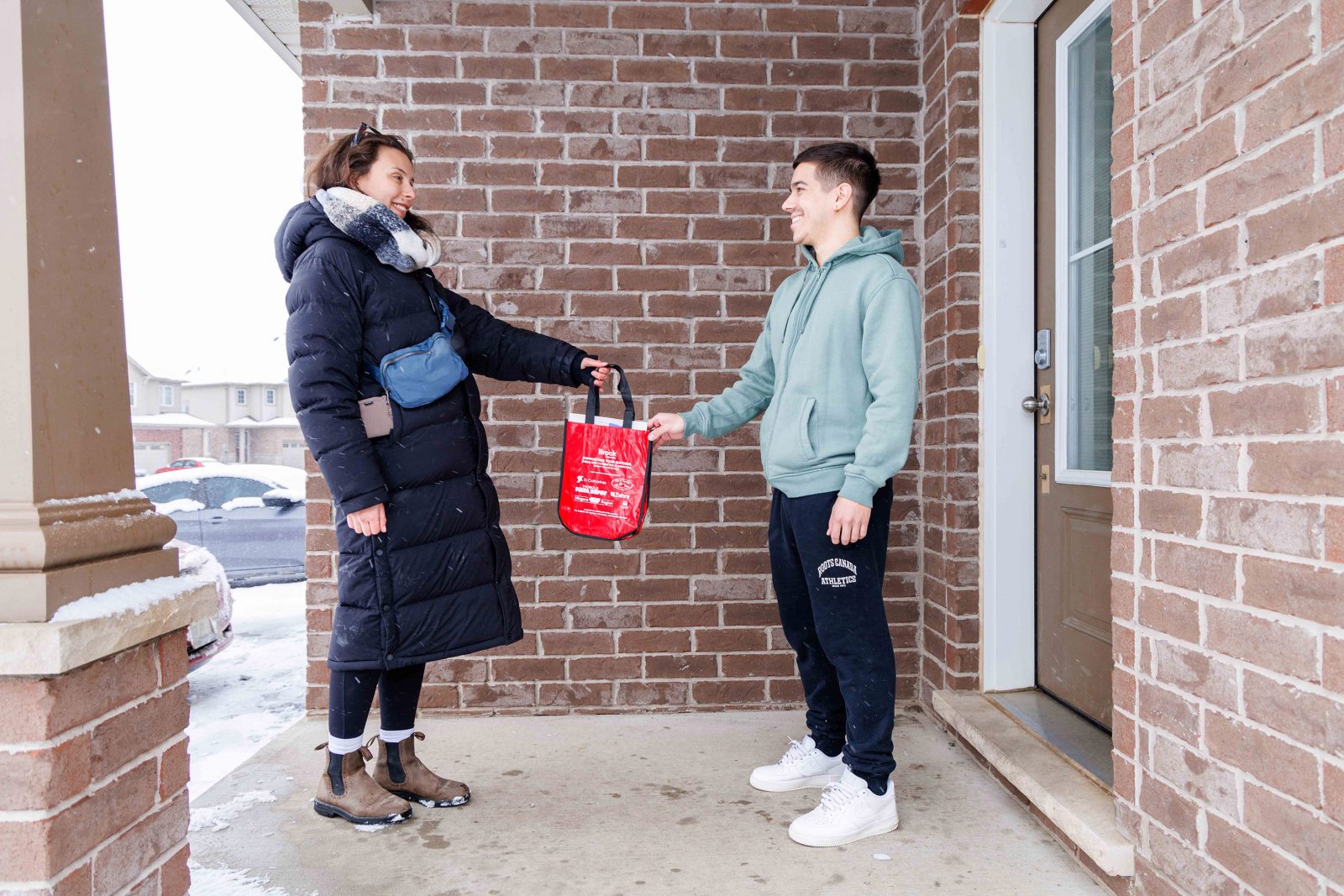 A woman hands a red back to a man on the front porch of a home.