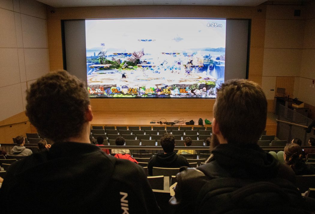 Two people sit in an auditorium space with a large screen displaying a video game.