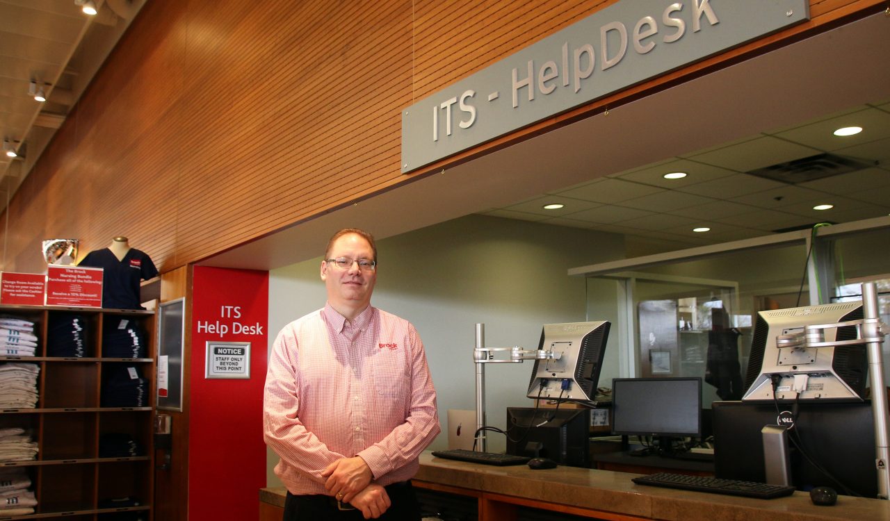 Gerald Cooper stands in front of a service desk with a computer on it. Above the desk is a sign that reads "ITS Help Desk".