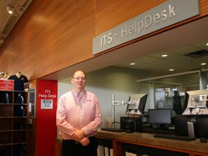 Gerald Cooper stands in front of a service desk with a computer on it. Above the desk is a sign that reads "ITS Help Desk".