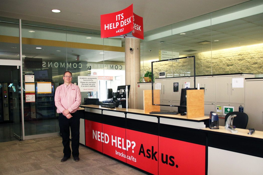 Gerald Cooper stands in front of a long service desk equipped with computers. A poster on the desk reads "Need help? Ask us." A sign above the desk reads "ITS Help Desk".